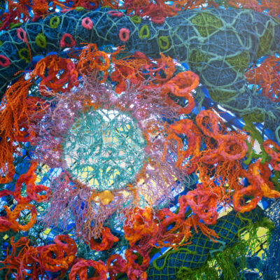 Sunlight shines through the woven recycled fishing nets on the vibrant and colorful abstract mural sculpture.