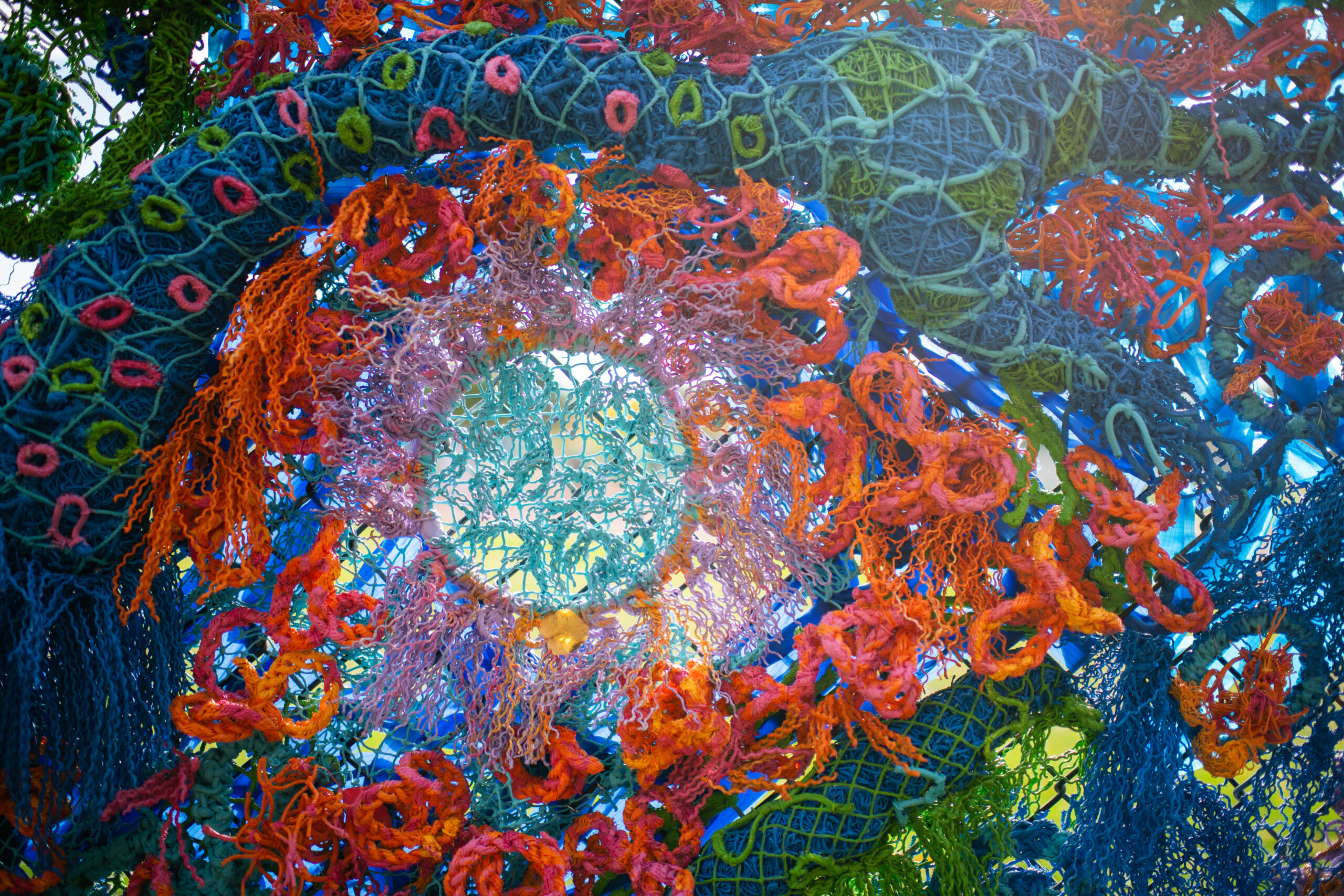 Sunlight shines through the woven recycled fishing nets on the vibrant and colorful abstract mural sculpture.